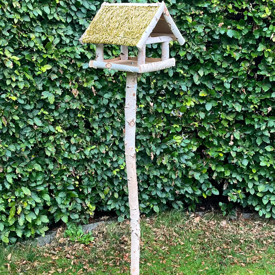 Fuglefoderhus i ægte birketræ med stråtag på stander (146 cm) fra HORTUS — rummeligt fuglehus hævet på spydstang, beskytter foder mod regn og sne og passer smukt i haven