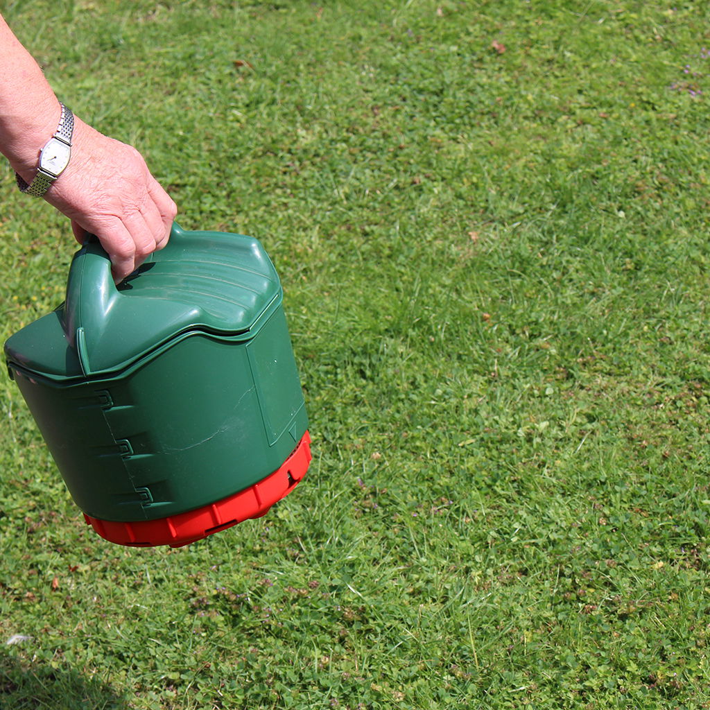 Person bærer grøn hånddrevet gødningsspreder på græsplæne