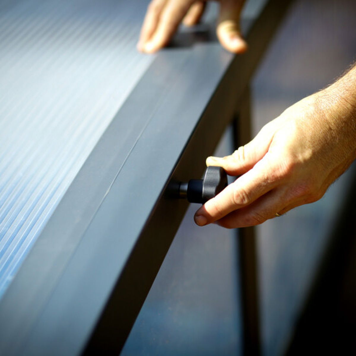 Hænder justerer låsemekanisme på terrasseoverdækning